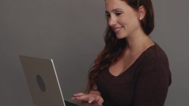 Gri arka planda dizüstü bilgisayar kullanan, gülümseyen, çilli genç bir kadının yakın görüntüsü. Eğitim ya da iş konsepti. Yüksek kalite 4k görüntü