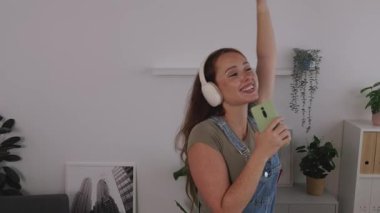 Genç ve mutlu bir kadın akıllı telefonunu mikrofon gibi tutarak şarkı söylüyor, evde neşeli bir enerji ve müzikle anın tadını çıkarıyor.