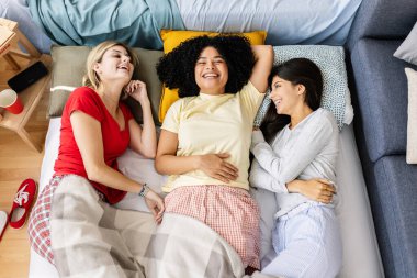 Pijama giyen üç neşeli genç kadın bir pijama partisinde yatakta uzanıyor, konuşuyor ve gülüyorlar. Kadın arkadaşlığı ve gençlik kavramı