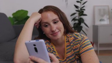 30 yaşında, yetişkin bir kadın rahat bir koltukta oturuyor, akıllı telefondan kayıyor ve internetten içeriği izliyor. Mobil teknoloji ve evde rahatlık ile rahat bir yaşam tarzı..