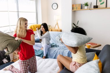 Multi-ethnic female friends enjoying a slumber party, playfully engaging in a pillow fight in pajamas. Female friendship and youth concept