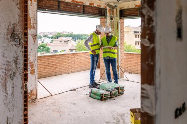 Two engineers wearing safety vests and helmets are using a tablet and evaluating building project progress at construction site. Engineering and architecture concept