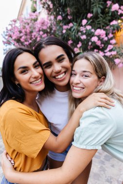 Three cheerful young women are hugging each other on a city street, enjoying their friendship and urban surroundings