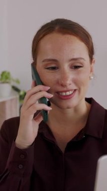 Vertical HD video of young successful adult businesswoman talking on phone while working on laptop computer at modern office. Professional female entrepreneur and business concept