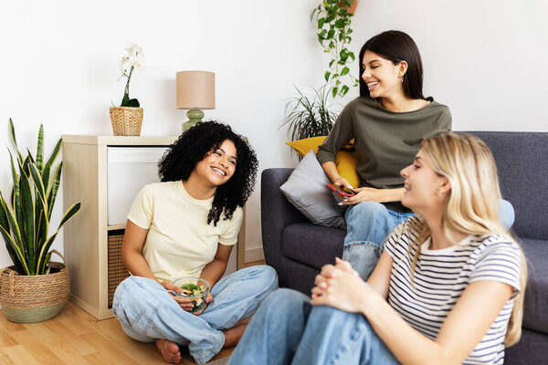 Three young women enjoying each others company, laughing and sharing a healthy salad while relaxing together in a cozy living room. Female friendship and youth concept