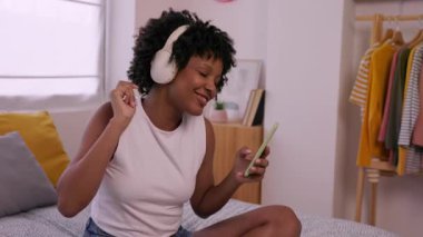Slow motion video of young happy African American woman sitting comfortably on her bed while wearing headphones and enjoying music on her smartphone. Relaxation, leisure and youth lifestyle concept.