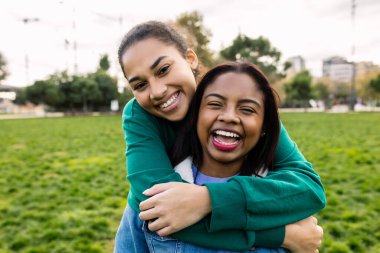 Close-up portrait of two Latin American teenage student girls sitting together at the park, sharing a joyful moment and smiling at the camera. Friendship, youth and outdoor lifestyle concept.