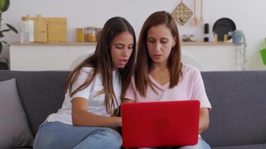 Young teenage student girl sitting on sofa at home with her mid adult mother, both sharing time together while using laptop computer. Family and technology lifestyle concept