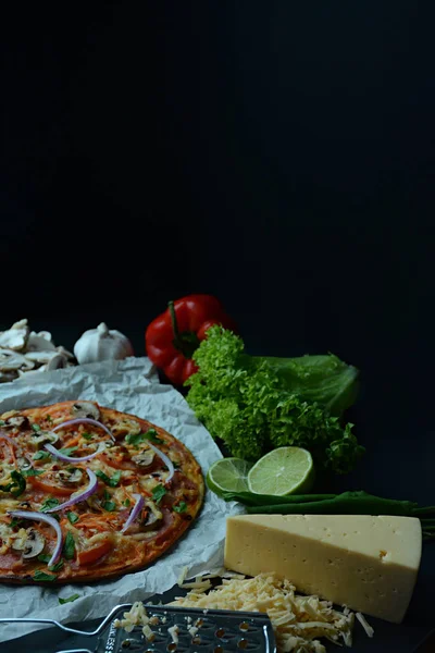 Fresh pizza with sausage, tomatoes, mushrooms and cheese on a dark background. Decorated with fresh vegetables and spices. Place for text. Side view.
