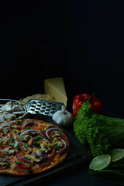 Fresh pizza with sausage, tomatoes, mushrooms and cheese on a dark background. Decorated with fresh vegetables and spices. Place for text. Side view.