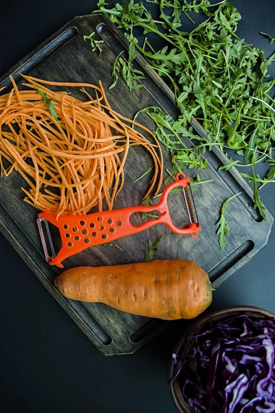 Cortar zanahorias en una tabla de roble. En el fondo cortando la col roja y rúcula sobre un fondo negro. Comida adecuada y saludable. Vista desde arriba . —  Fotos de Stock
