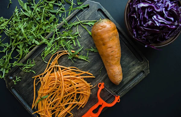 Cortar zanahorias en una tabla de roble. En el fondo cortando la col roja y rúcula sobre un fondo negro. Comida adecuada y saludable. Vista desde arriba . —  Fotos de Stock