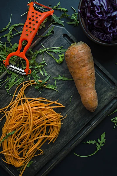 Cortar zanahorias en una tabla de roble. En el fondo cortando la col roja y rúcula sobre un fondo negro. Comida adecuada y saludable. Vista desde arriba . —  Fotos de Stock