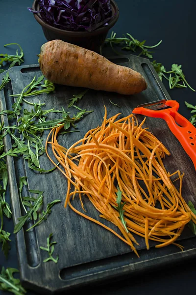 Нарезание моркови на дубовой доске. На заднем плане режут красную капусту и рукколу на черном фоне. Правильное и здоровое питание . — стоковое фото