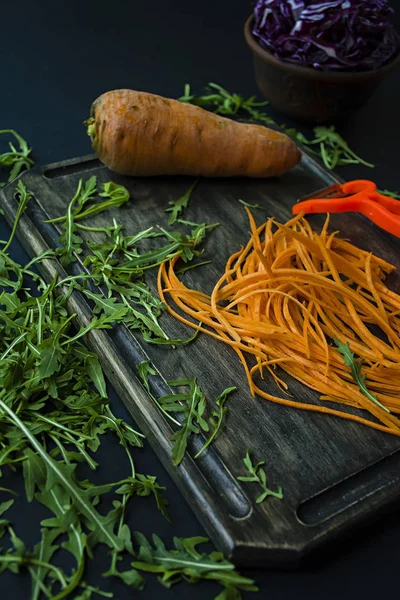 Нарезание моркови на дубовой доске. На заднем плане режут красную капусту и рукколу на черном фоне. Правильное и здоровое питание . — стоковое фото