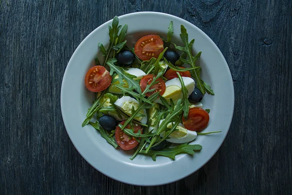 Görög saláta friss paradicsommal, arugula, tojás, olajbogyó, olívaolajjal, sötét fa háttérrel. Egészséges táplálkozás. Vega étel. — Stock Fotó