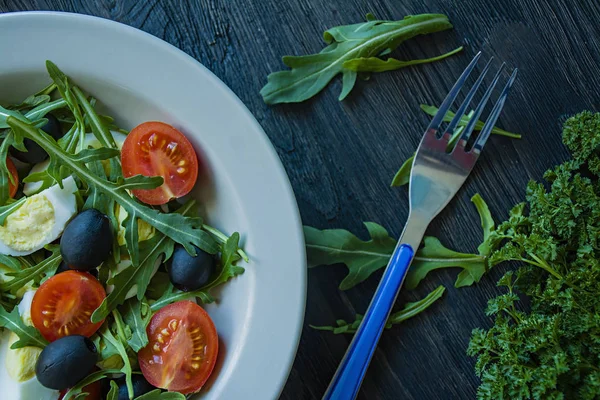 Görög saláta friss paradicsommal, arugula, tojás, olajbogyó, olívaolajjal, sötét fa háttérrel. Egészséges táplálkozás. Vega étel. — Stock Fotó