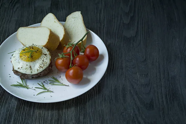 Marha steak tükörtojással fűszerekkel. Rozmaringos, friss Cseresznyés és kenyérszelet díszítve. Benyújtani egy fehér lemez. Sötét fából készült háttér. Oldalnézetből. Szóköz a szöveghez. — Stock Fotó