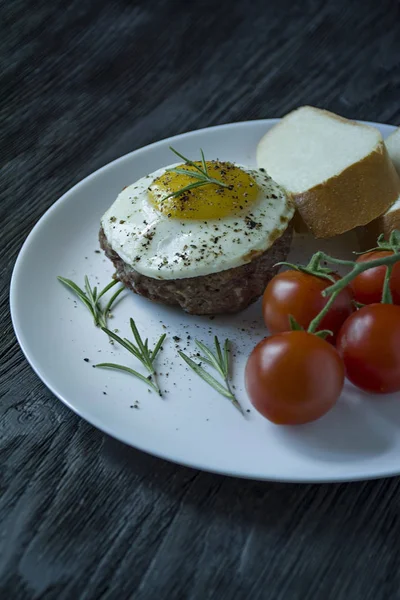 Marha steak tükörtojással fűszerekkel. Rozmaringos, friss Cseresznyés és kenyérszelet díszítve. Benyújtani egy fehér lemez. Sötét fából készült háttér. Oldalnézet. — Stock Fotó