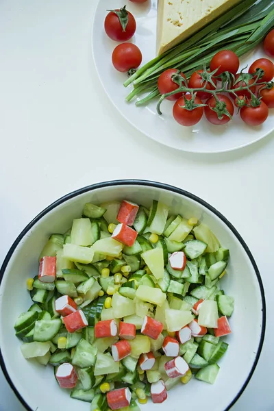 Ensalada con maíz, palitos de cangrejo, pepinos en un bol blanco sobre un fondo blanco. Ensalada vegetariana. Proceso de cocción . —  Fotos de Stock