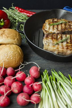 Siyah ahşap arka planda et, biberiye, yeşillikler ve sebzeler için baharatlar ile süslenmiş yuvarlak ızgara tavada ızgara biftek.