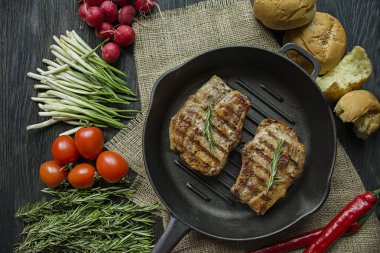 Siyah ahşap arka planda et, biberiye, yeşillikler ve sebzeler için baharatlar ile süslenmiş yuvarlak ızgara tavada ızgara biftek.