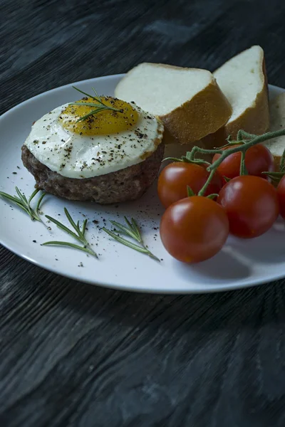 Marha steak tükörtojással fűszerekkel. Rozmaringos, friss Cseresznyés és kenyérszelet díszítve. Benyújtani egy fehér lemez. Sötét fából készült háttér. Oldalnézetből. Szóköz a szöveghez. — Stock Fotó