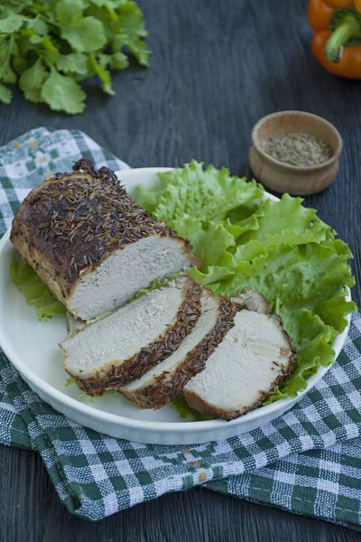 Baked pork tenderloin in spices sliced on a white plate with green salad. Dark wooden background.
