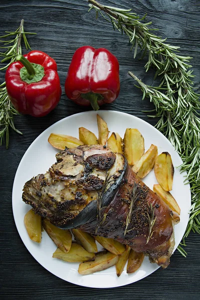 Fried pork knuckle with potatoes served on a white plate. Decorated with fresh Bulgarian pepper, rosemary. Dark wooden background. View from above. — Stock Photo, Image