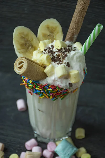 Banana milkshake with ice cream and whipped cream, marshmallows, cookies, waffles, served in a glass cup decorated with confectionery dressing. Dark wooden background.
