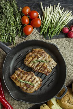 Siyah ahşap arka planda et, biberiye, yeşillikler ve sebzeler için baharatlar ile süslenmiş yuvarlak ızgara tavada ızgara biftek.