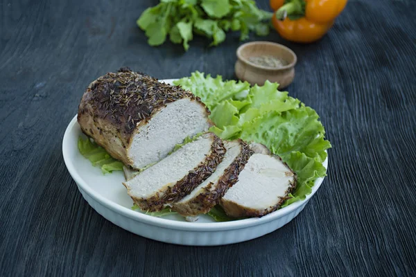Baked pork tenderloin in spices sliced on a white plate with green salad. Dark wooden background.