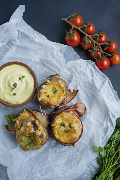 Запеченный новый картофель, завернутый в бекон, с сыром, соусом и свежими овощами. Темный фон — стоковое фото