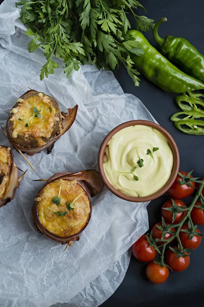 Запеченный новый картофель, завернутый в бекон, с сыром, соусом и свежими овощами. Темный фон . — стоковое фото