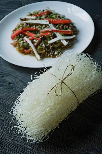 Asian cuisine. Raw cellophane noodles. In the background is decorated with cellophane noodles salad with vegetables, greens. Funchoza. Proper nutrition. Healthy food. View from above. Dark wooden back