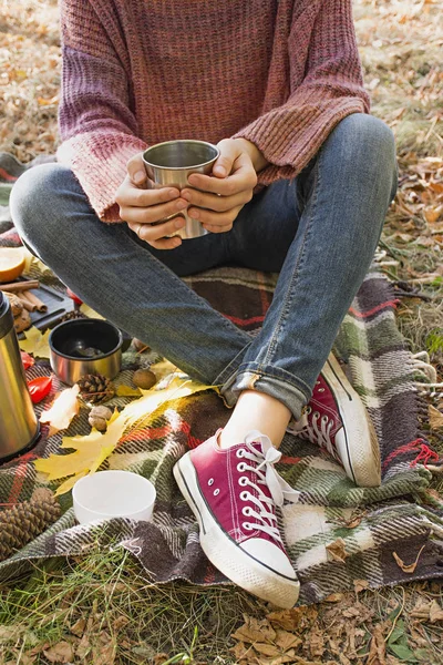 Autumn picnic in the park. The girl holds a cup of tea in her hands. Basket with flowers on a blanket in yellow autumn leaves. Autumn concept.