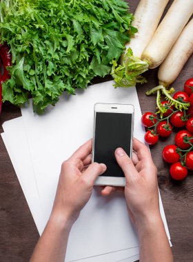 Erkek eller mutfak masasındaki akıllı telefonu taze sebzelerin arka planına karşı tutarlar. Yemek arkaplanı. Sebze geçmişi. Süper yemek. Düz yatıyordu. Boşluğu kopyala.