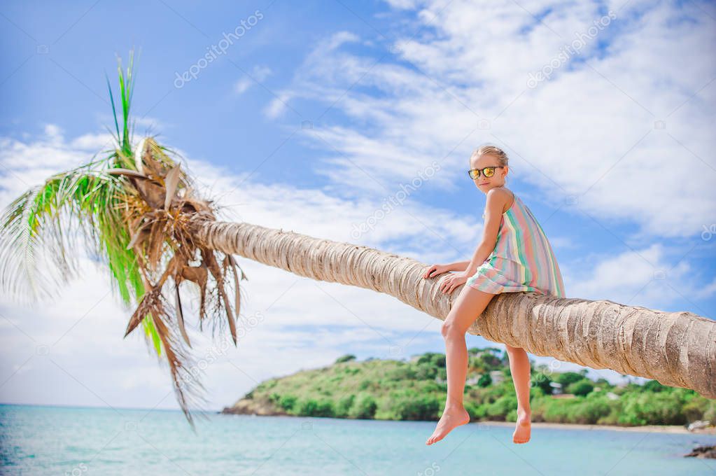 Adorable little girl sitting on palm tree during summer vacation on white beach