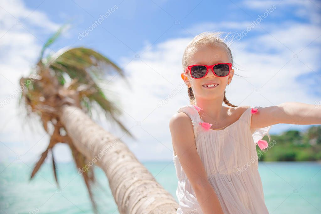 Adorable little girl sitting on palm tree during summer vacation on white beach