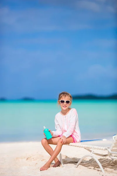 Little girl with bottle of sun cream sitting at tropical beach — Stock Photo, Image