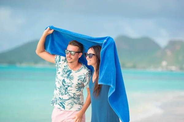 Pareja joven en la playa tropical bajo la toalla escondida de la lluvia tropical . — Foto de Stock