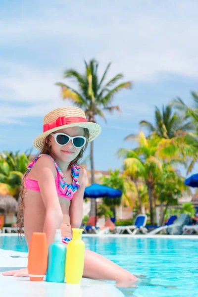 Little girl with bottle of sun cream sitting on the edge of swimming pool — Stock Photo, Image