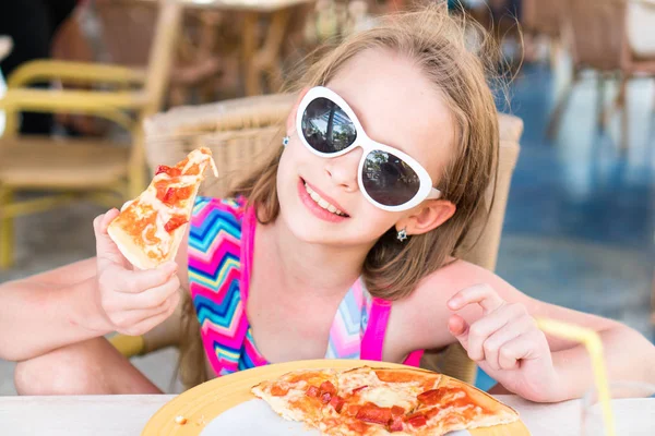 Retrato de una linda niña sentada junto a la mesa y comiendo pizza —  Fotos de Stock