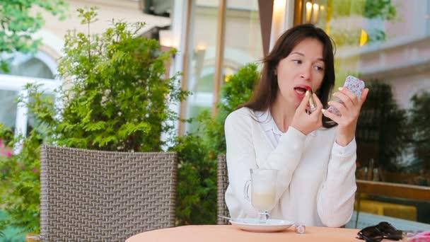 Belle femme peignant ses lèvres avec du rouge à lèvres assis dans un café extérieur. Jeune fille avec délicieux cappuccino sucré . — Video