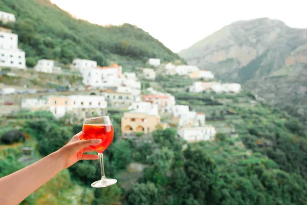 Weibliche Hand hält Glas mit Spritz Aperol Alkohol trinken Hintergrund der schönen alten italienischen Dorf an der Amalfi-Küste — Stockfoto
