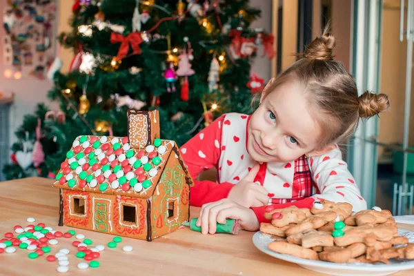 Kleine Mädchen basteln Weihnachts-Lebkuchenhaus am Kamin im geschmückten Wohnzimmer. — Stockfoto