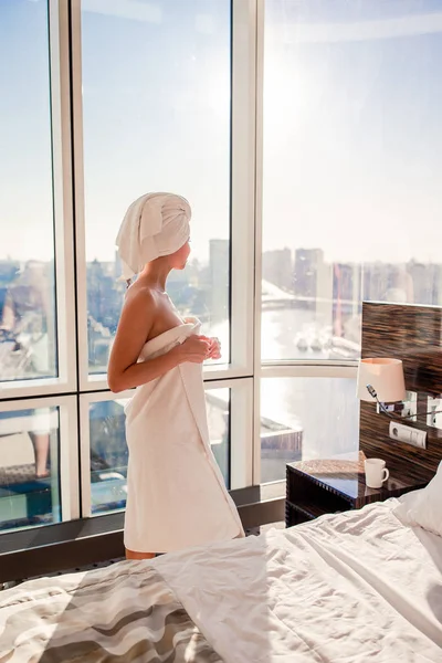 Schöne junge Frau in weißem Badetuch auf dem Kopf entspannt sich nach der Dusche auf dem Bett mit einer Tasse Kaffee — Stockfoto