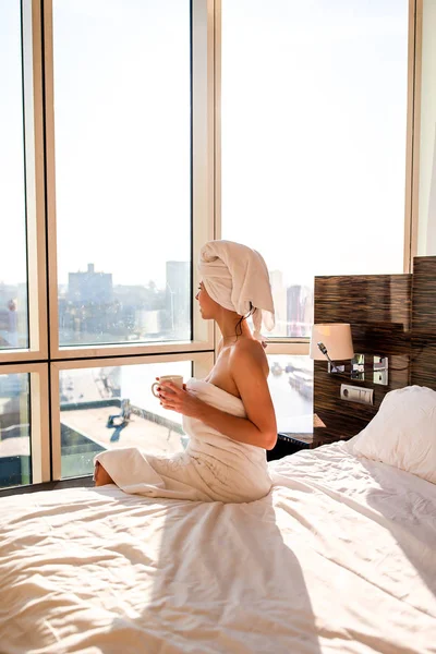 Hermosa mujer joven en toalla de baño blanca en la cabeza relajante después de la ducha en la cama con taza de café —  Fotos de Stock