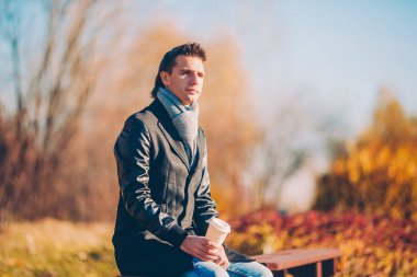 Genç adam sonbahar parkında telefonla kahve içiyor.