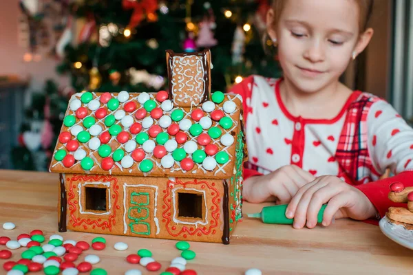 Kleine Mädchen basteln Weihnachts-Lebkuchenhaus am Kamin im geschmückten Wohnzimmer. — Stockfoto